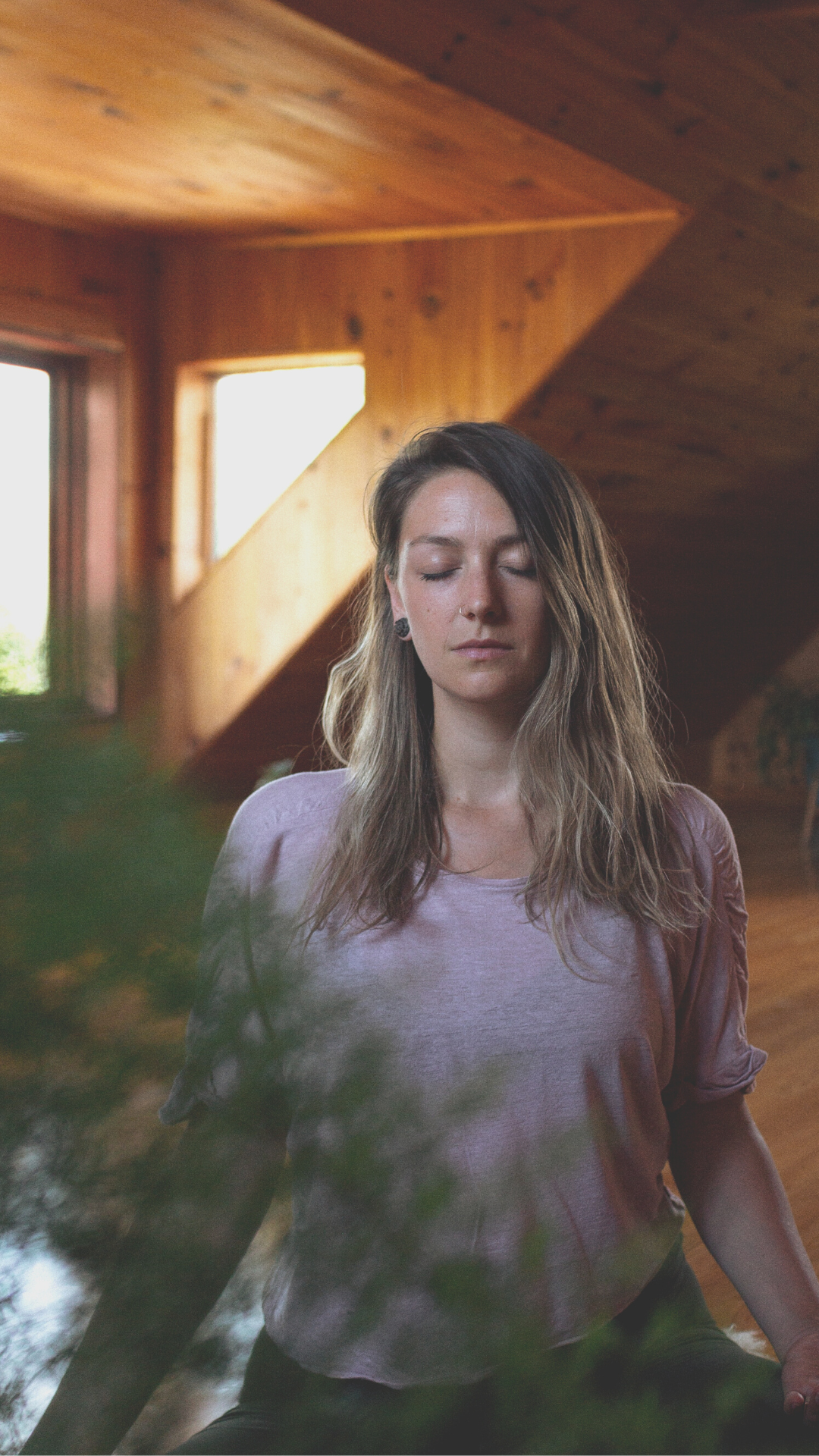 Naomi Elder sitting meditating with eyes closed in a light pink shirt. studio in background has wood walls and windows. blurry green plant in left corn of the foreground.